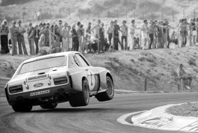 Rear-three-quarter view of a white Ford touring car cresting a corner on the Nordschleife with spectators packed behind the fence.