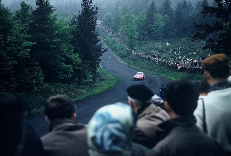 Spectators line a misty forest stretch of the Nordschleife as a small saloon car climbs the hill, Eifel region, mid-1960s.