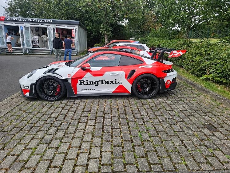 A red-and-white Porsche 911 GT3 RS in Ring Taxi livery parked on cobblestones outside the Official Co-Pilot Partner kiosk at the Nürburgring.