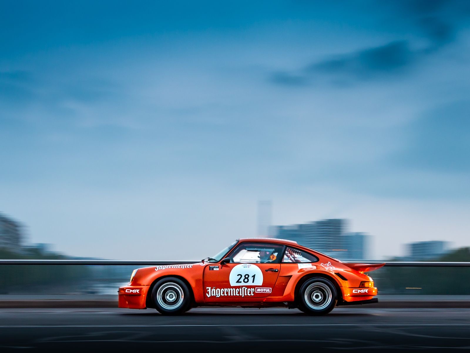 Orange Jagermeister Porsche 911 RSR race car at speed across a city bridge at dusk